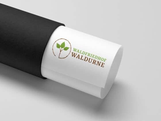 Zusammengerolltes weißes Papier mit grün-braunem "Waldfriedhof Waldurne"-Logo teils in schwarzer Röhre. Zusammengerolltes weißes Papier mit grün-braunem "Waldfriedhof Waldurne"-Logo teils in schwarzer Röhre.